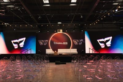Main room at The Whalies 2026 with a stage before a glowing circular backdrop, large screens and rows of clear chairs