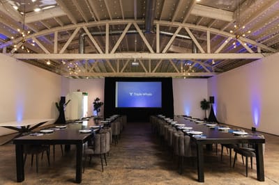 Empty boardroom with two long black tables, grey chairs and a Triple Whale screen under twinkle lights