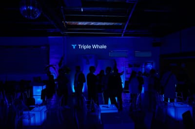 Guests in silhouette under Triple Whale lettering in a dim blue room with glowing cube tables and a disco ball