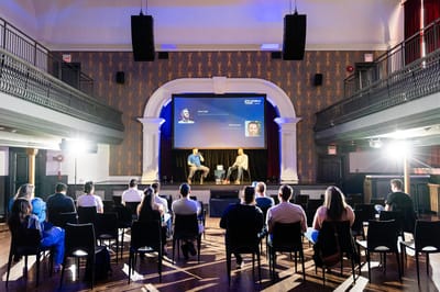 Panel on stage under a DTC World Tour screen in a hall with patterned wallpaper, balconies and a seated audience
