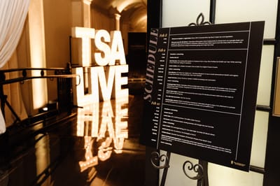 Lit TSA LIVE marquee letters reflected on a glossy floor beside a schedule board on an easel