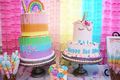 Rainbow-striped Khloe cake and a white unicorn cake reading Happy 1st Birthday in front of pastel paper garlands