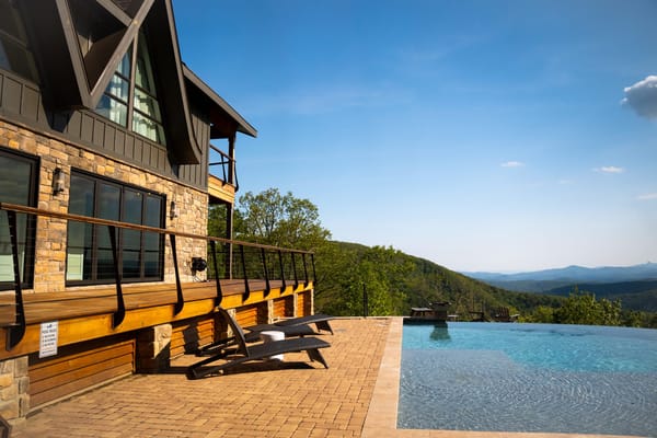 Stone house with a wooden deck, lounge chairs and an infinity pool facing blue mountains