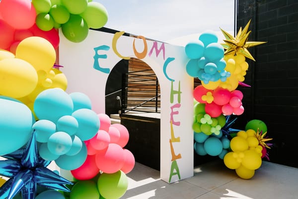 Empty white arched ECOMCHELLA entrance with teal, pink, yellow and green balloons and blue star balloons