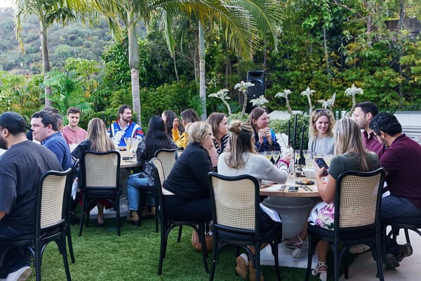 Guests at round wooden tables with cane-back chairs, orchids and black candles on a lawn below the hills