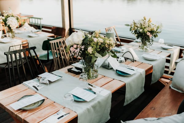 Wooden tables with sage runners, blue plates, menu cards and vases of flowers beside the water