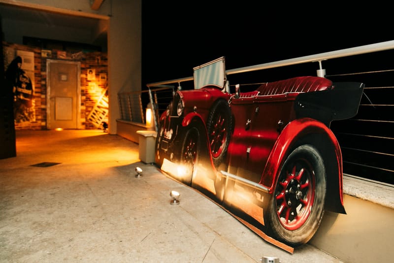 Flat cutout of a red vintage car along a railing at night, with a brick speakeasy front behind