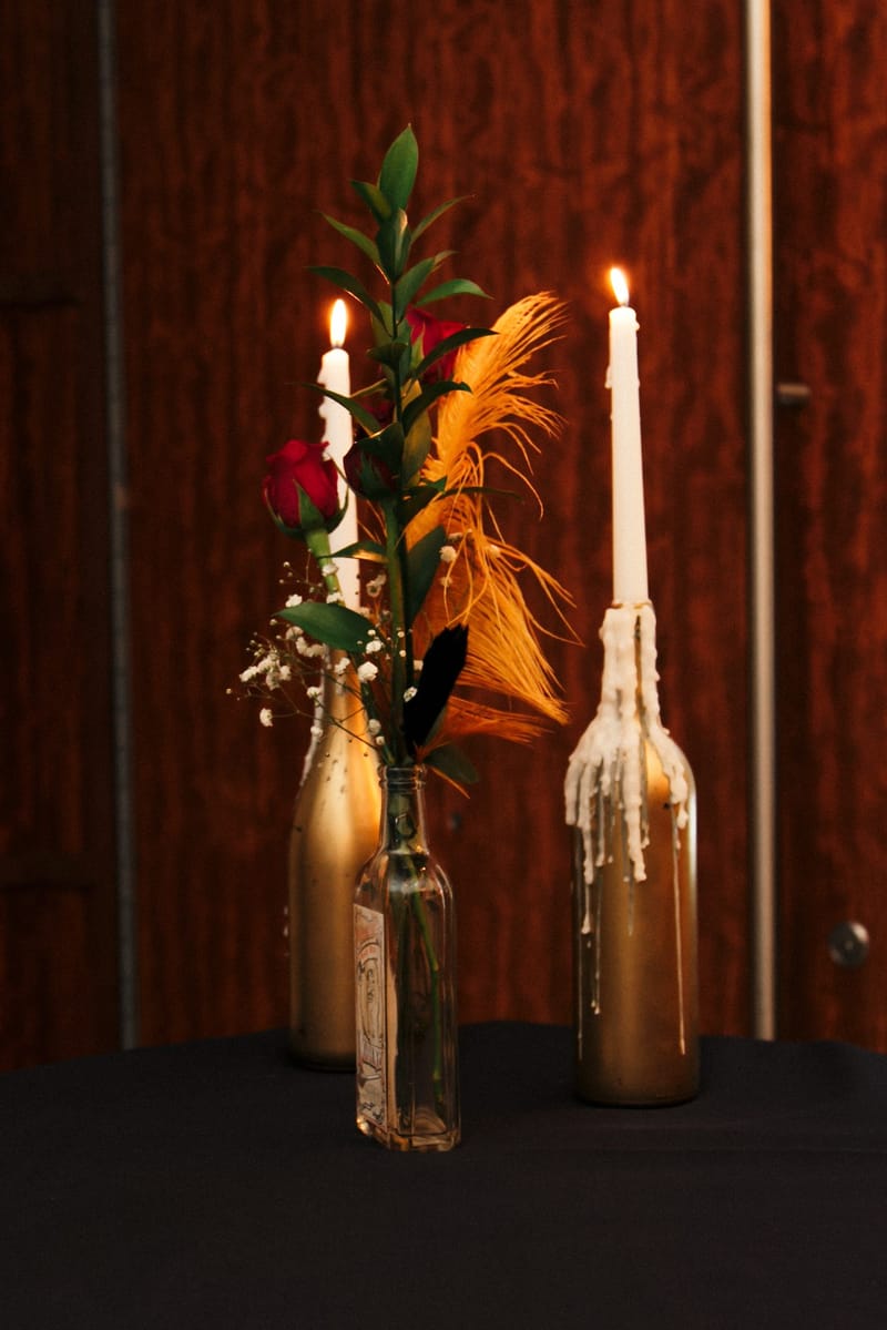 Taper candles dripping wax in gold-painted bottles beside a red rose and a gold feather