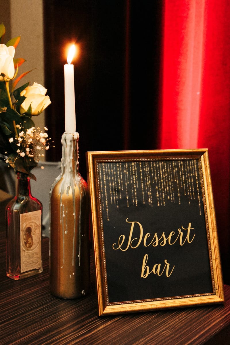 Gold-framed Dessert bar sign beside a gold-painted bottle holding a lit candle