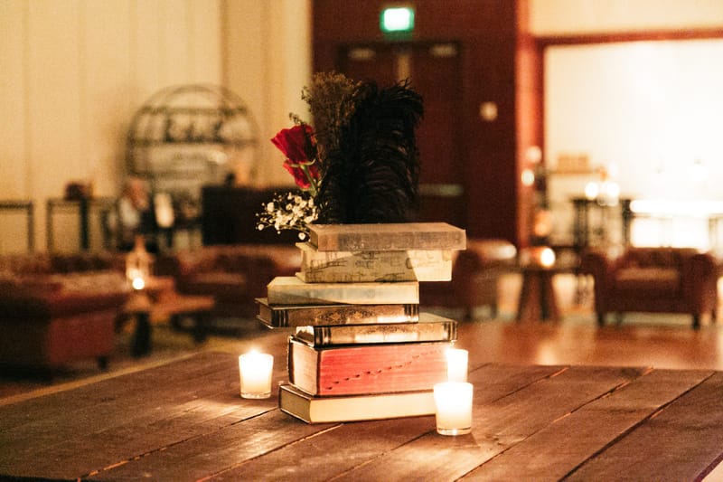 Stack of old books with a black feather and red flowers between candles, leather chairs blurred behind