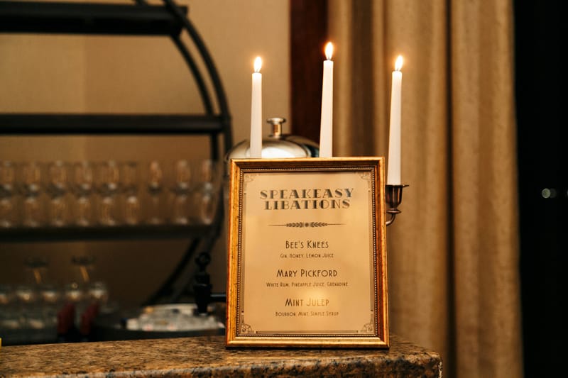 Gold-framed Speakeasy Libations menu listing Bee's Knees, Mary Pickford and Mint Julep, with candles behind