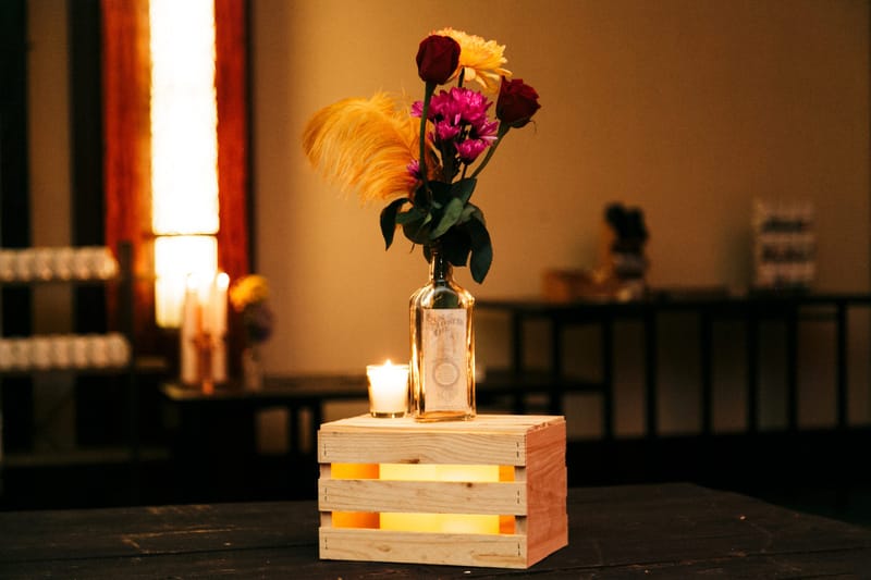 Lit wooden crate topped with a bottle of red and pink roses, a feather and a candle