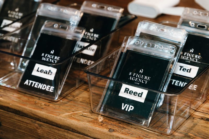 Clear bins of 8 Figure Agency Live name badges marked ATTENDEE and VIP on a wooden table
