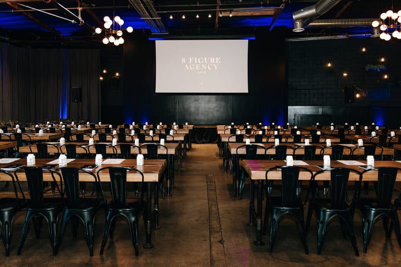 Rows of wooden tables and black metal chairs facing an 8 Figure Agency Live screen