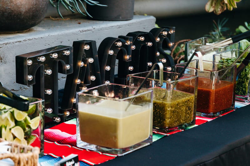 Light-up TACOS marquee letters behind glass dishes of salsas and limes on a striped runner