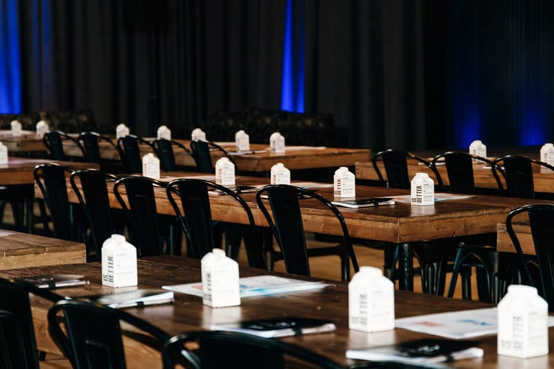 Wooden tables set with notebooks, papers and boxed water cartons in front of black chairs