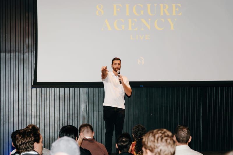 Speaker with a microphone pointing at the audience under an 8 Figure Agency Live screen
