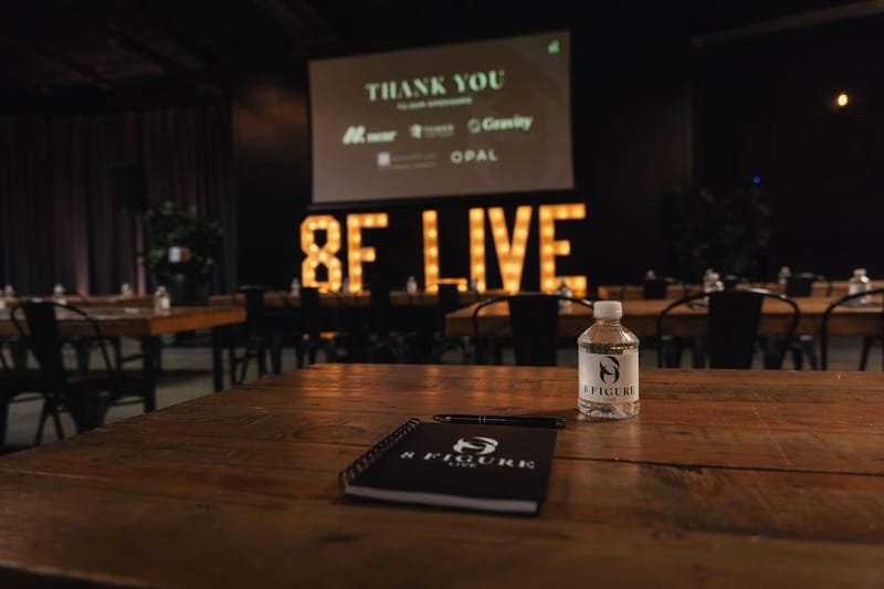 An 8 Figure notebook and a water bottle on a wooden table, with the lit 8F LIVE letters behind