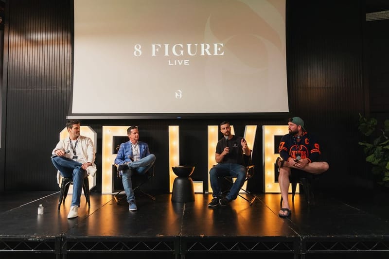 Four panelists seated on stage under an 8 Figure Live screen, with the lit letters behind them