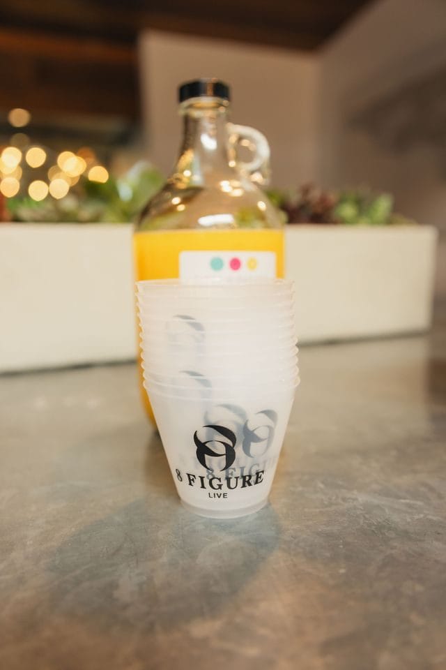 Glass jug of orange drink behind a stack of cups printed with the 8 Figure Live logo