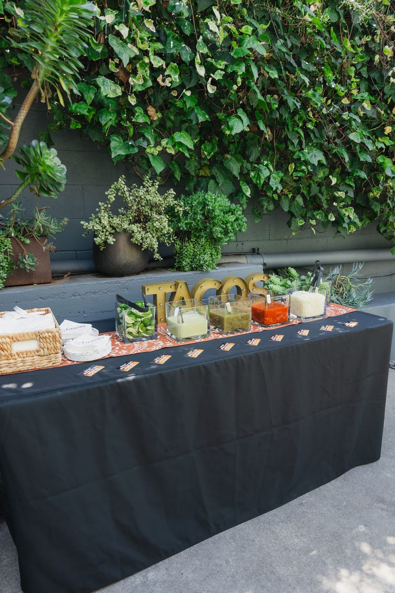 A taco bar under an ivy wall, with gold TACOS letters, glass bowls of limes and salsas, and a basket of plates