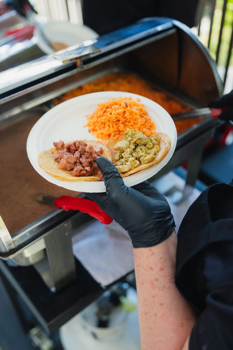 A server in black gloves holding a plate with two tacos and rice over a chafing dish