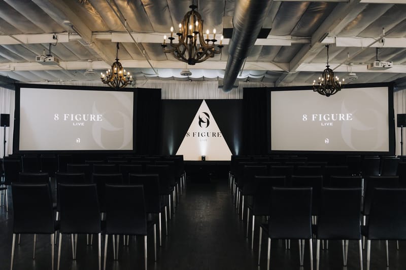 Rows of black chairs facing two 8 Figure Live screens and a white triangle stage backdrop under chandeliers