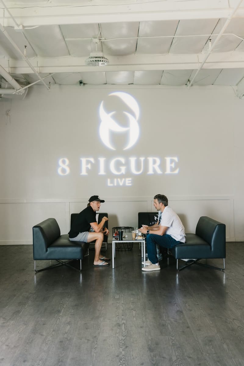 Two guests talking on black sofas beneath the 8 Figure Live logo projected on a white wall