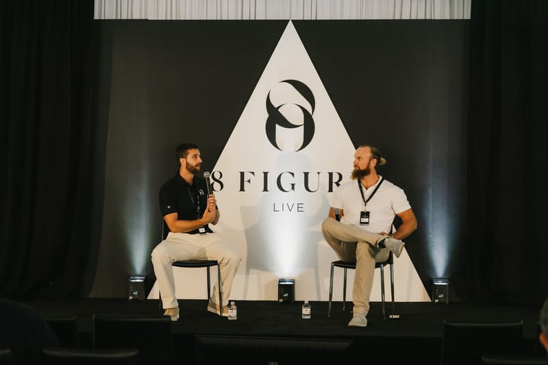 Two speakers in a fireside chat on stools in front of the white 8 Figure Live triangle