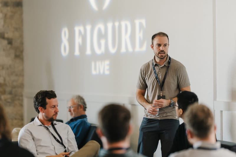 Speaker holding a microphone in front of the projected 8 Figure Live logo while attendees listen