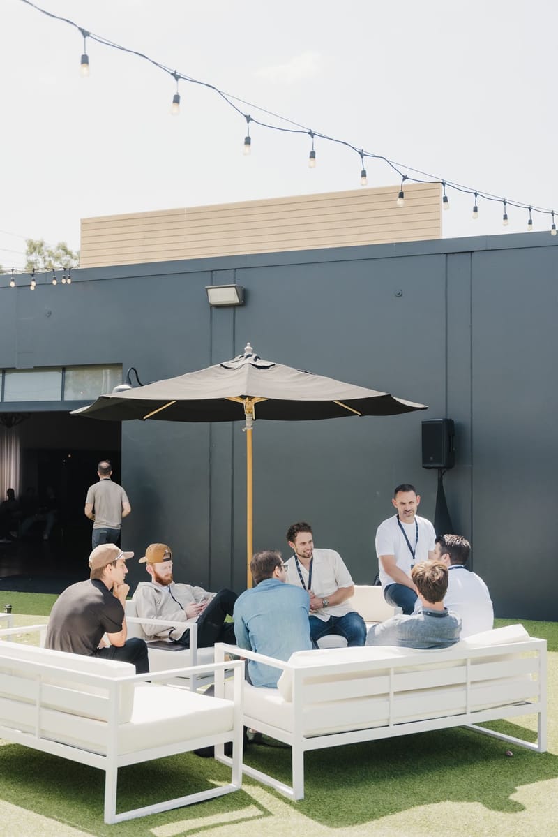 Attendees on white outdoor sofas under a black umbrella on the turf patio