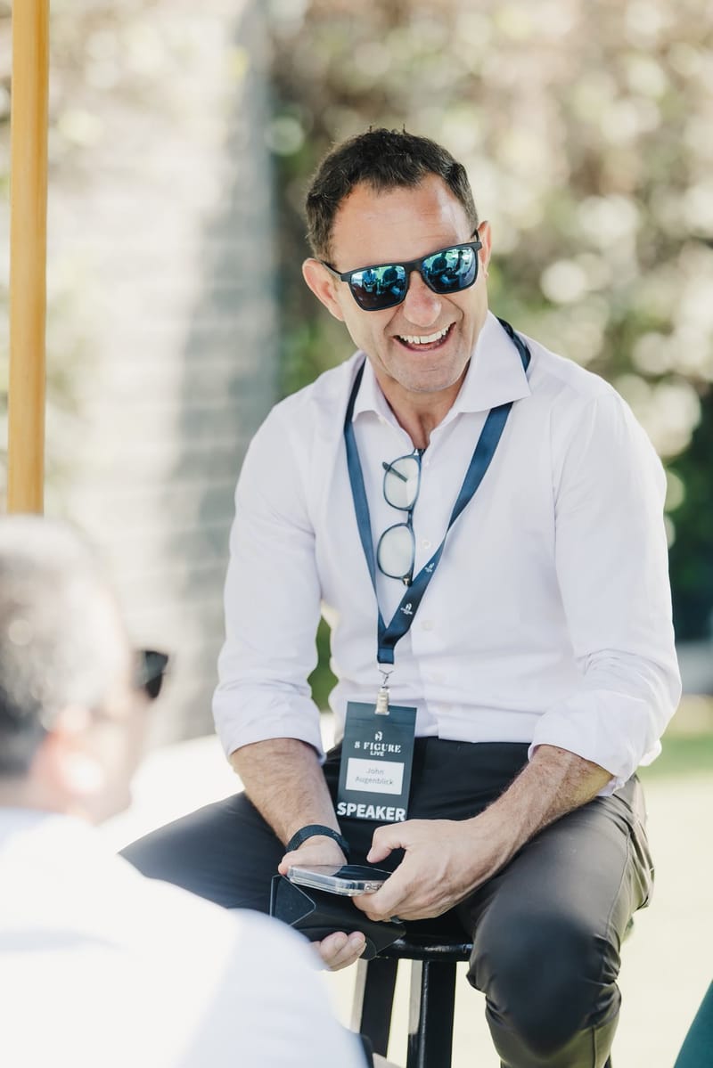 Speaker in sunglasses laughing on a stool outdoors