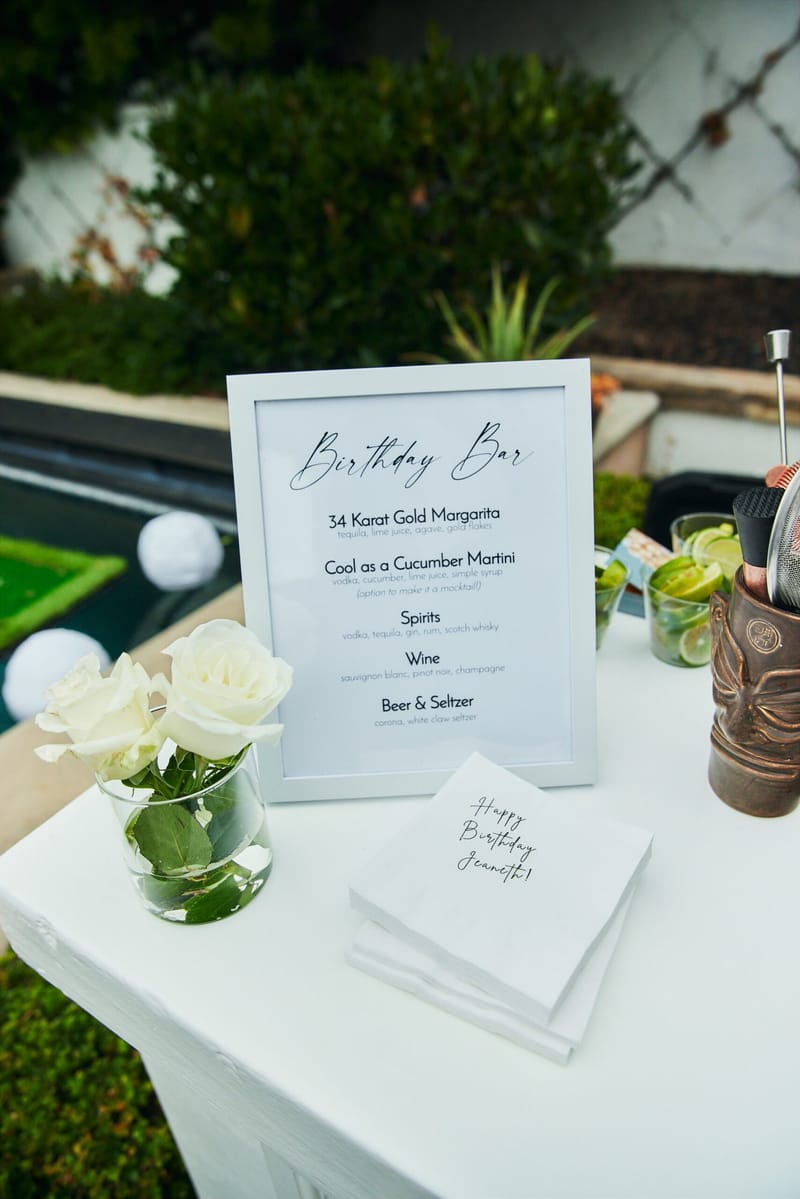 Birthday Bar menu listing a 34 Karat Gold Margarita beside napkins printed Happy Birthday Jeaneth! and a white rose