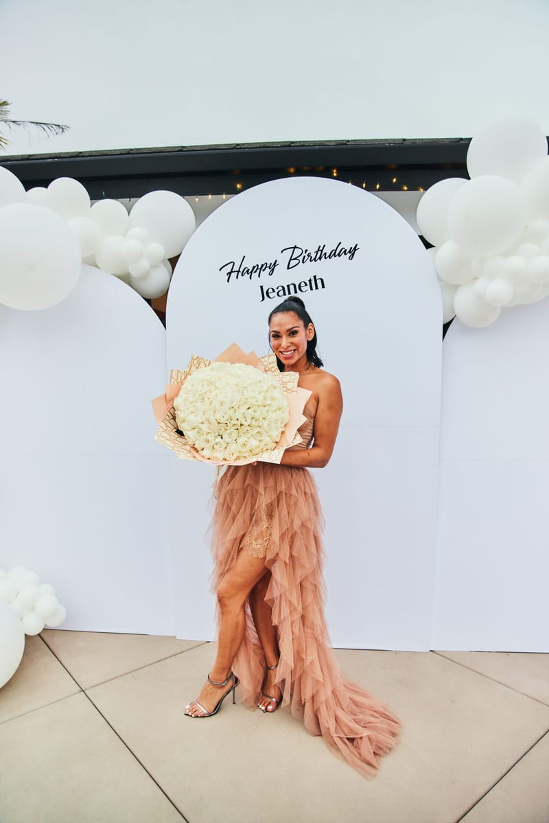 Guest of honor in a blush tulle gown holding white roses before white arched panels reading Happy Birthday Jeaneth