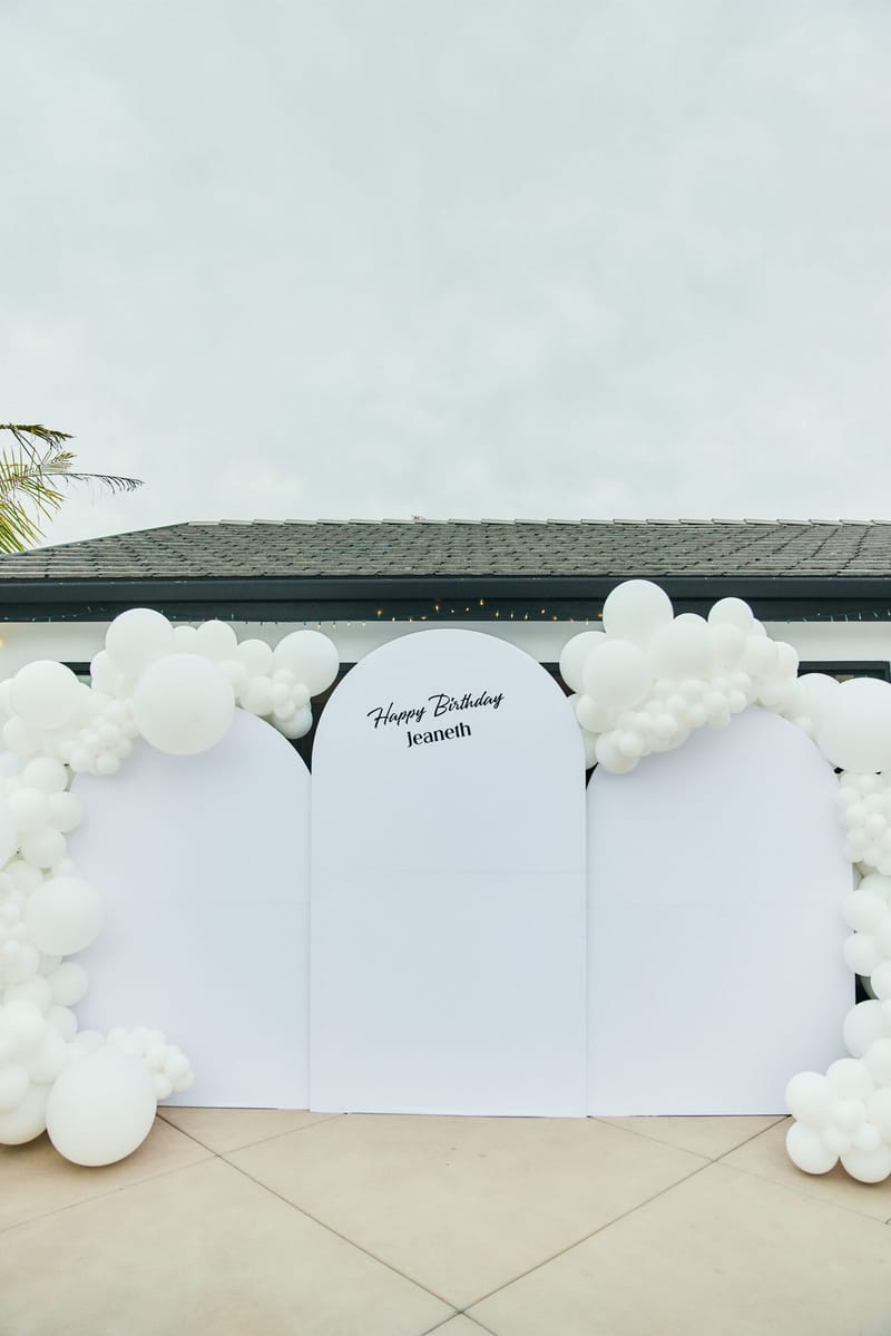 White arched panels reading Happy Birthday Jeaneth framed by clusters of white balloons on a patio