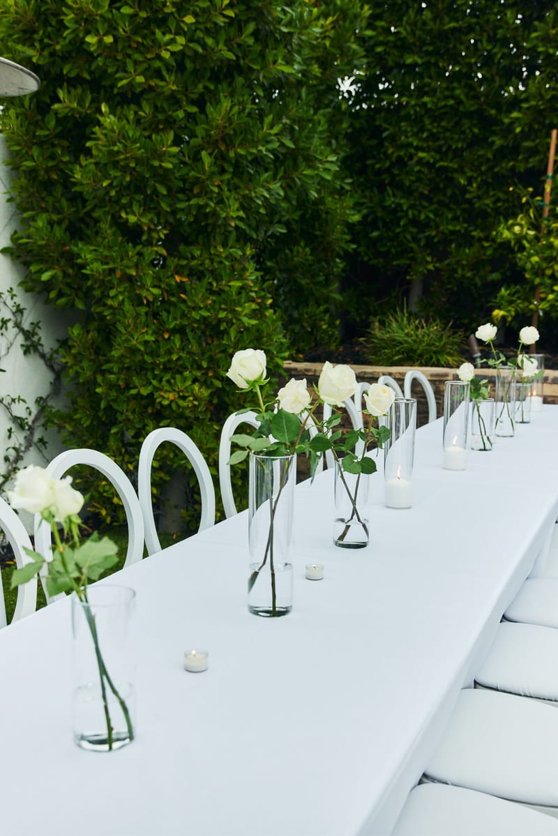 Long white table with white roses in tall glass vases, small candles and white loop-back chairs by a hedge