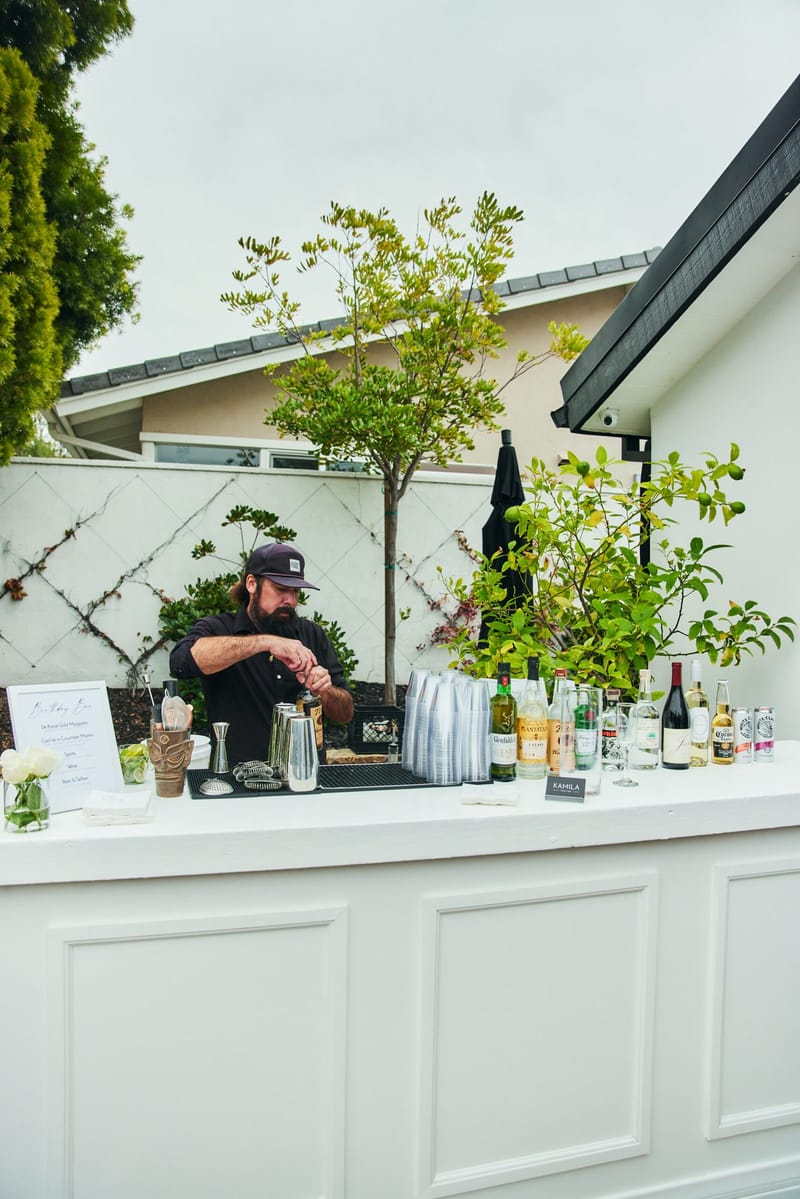 Bartender mixing drinks at a white paneled bar with the Birthday Bar menu, bottles and stacked cups