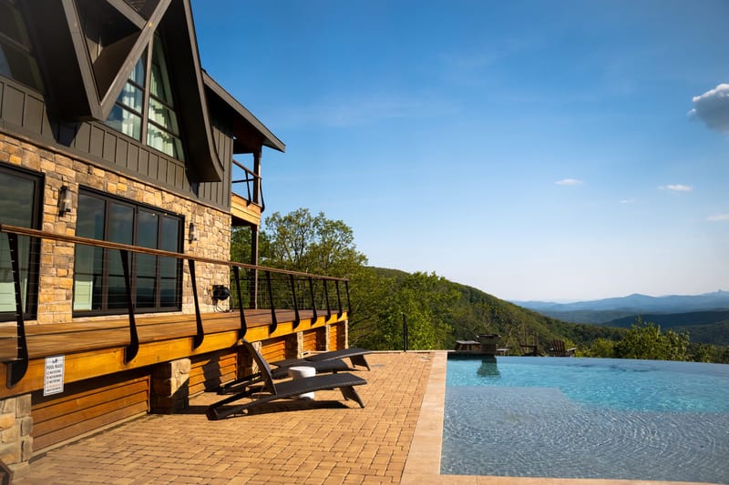Stone house with a wooden deck, lounge chairs and an infinity pool facing blue mountains