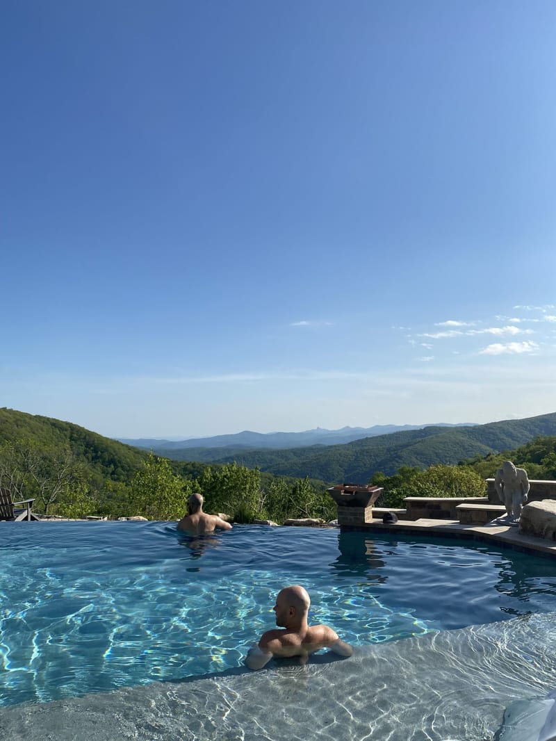 Two guests in an infinity pool looking out over layers of green mountains under a clear sky