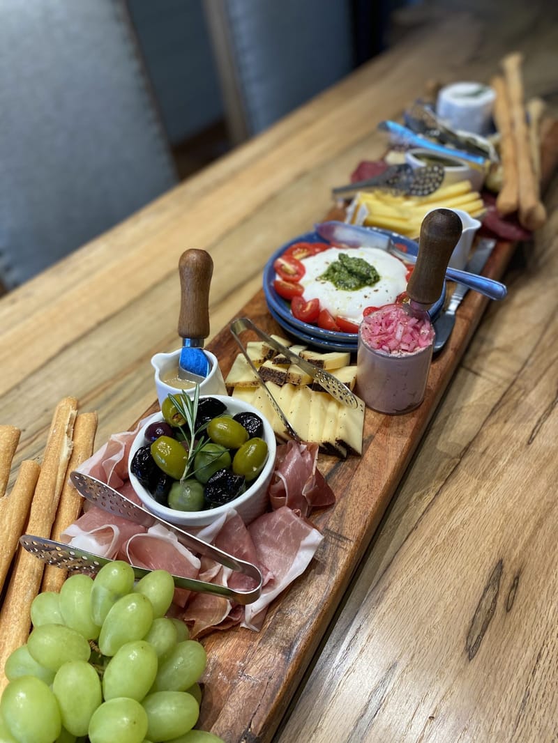 Long wooden board of cured meats, cheese, burrata with pesto, olives and green grapes