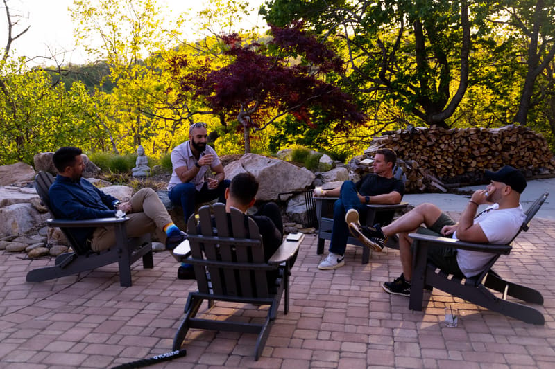 Guests talking in dark Adirondack chairs on a brick patio beside a stack of firewood