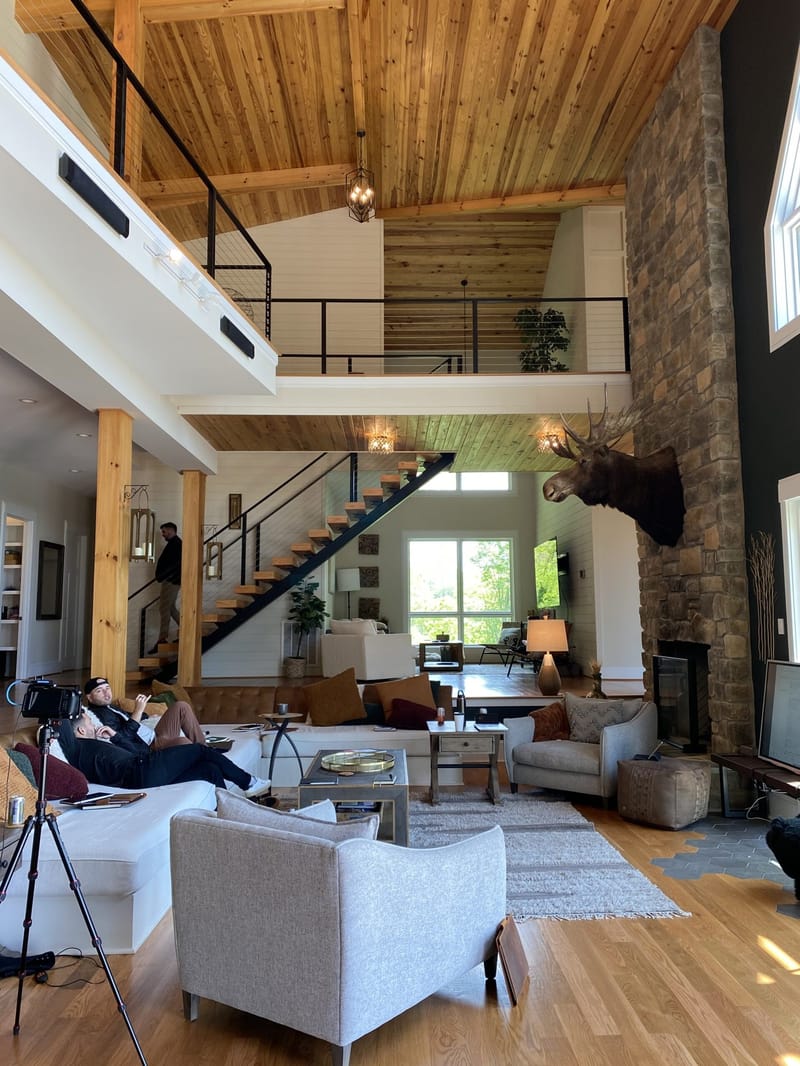 Great room with a timber ceiling, stone fireplace wall, open staircase and a camera on a tripod
