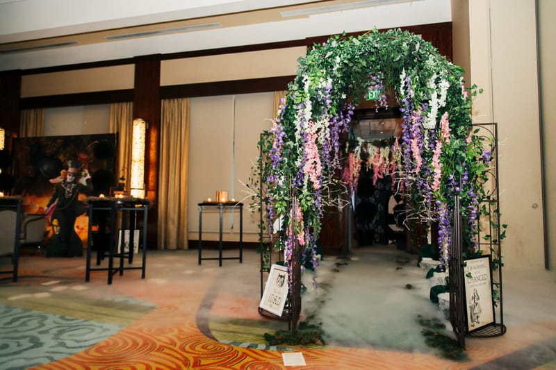 Entrance trellis hung with purple, pink and white flowers over low fog, with a Mad Hatter cutout to one side