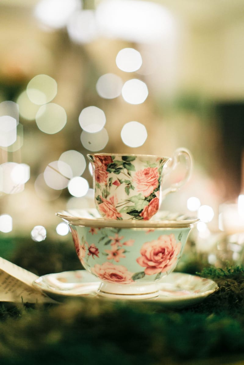 Two floral teacups stacked on saucers on moss, with fairy lights glowing behind