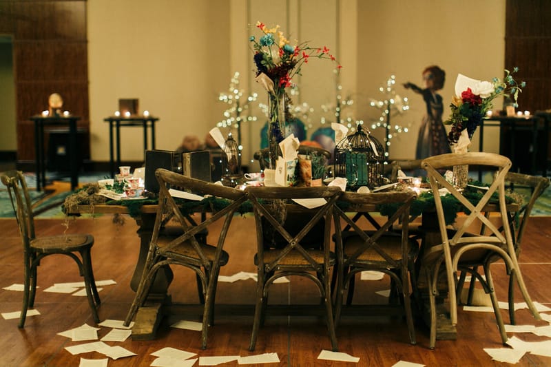 Long wooden table with cross-back chairs, teacups, birdcages and flowers, loose book pages scattered on the floor