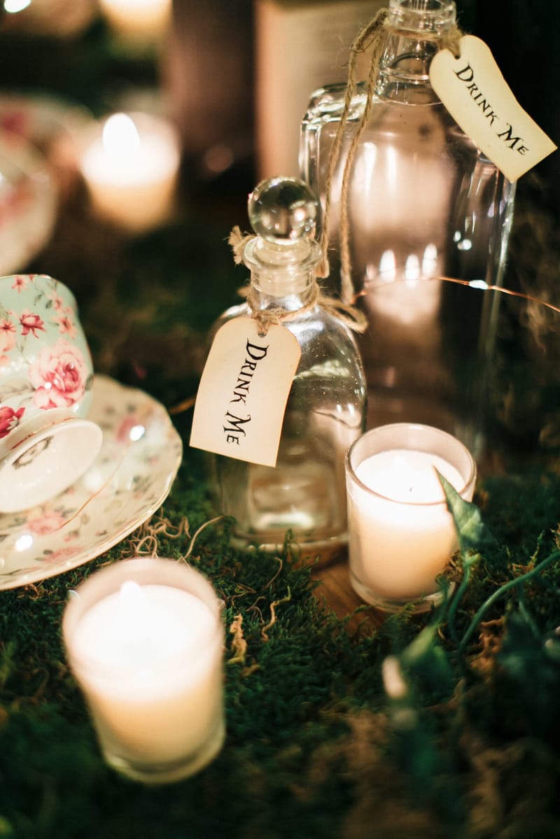 Glass bottles with Drink Me tags among votive candles and a floral teacup on moss