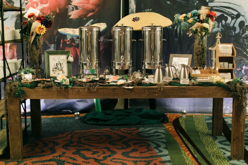 Tea station with three silver urns, tea bags and flowers on a moss-trimmed rustic table before a painted mural