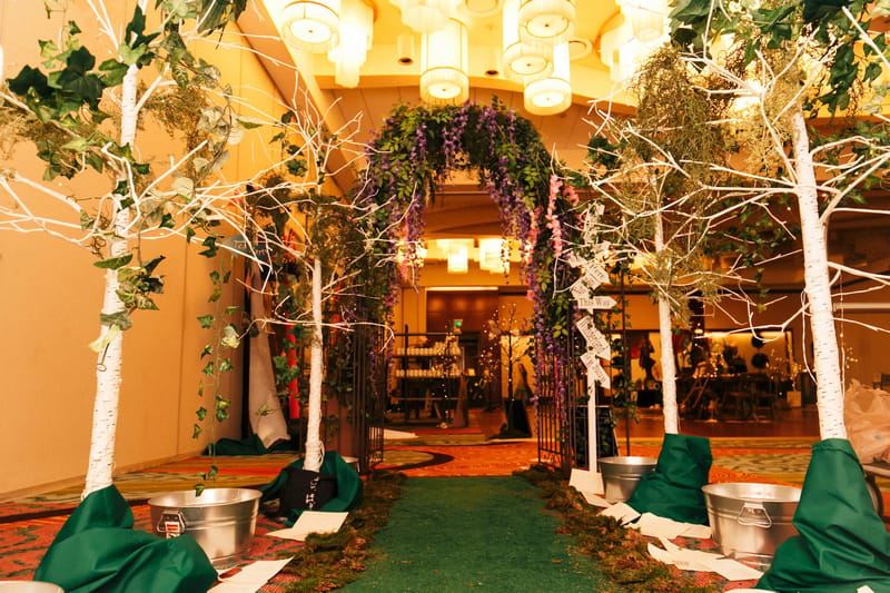 Green runner between white birch trees and galvanized tubs leading to a doorway hung with purple flowers
