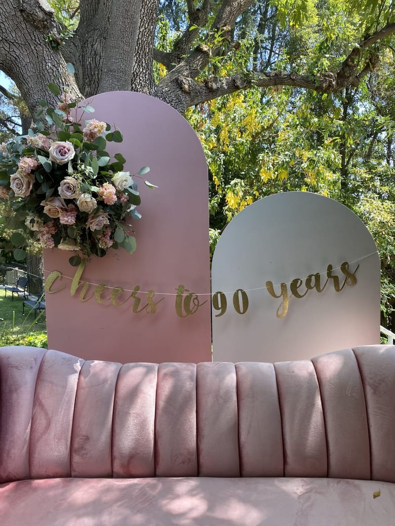 Pink channel-tufted velvet sofa in front of two tall rounded panels hung with a gold cheers to 90 years banner