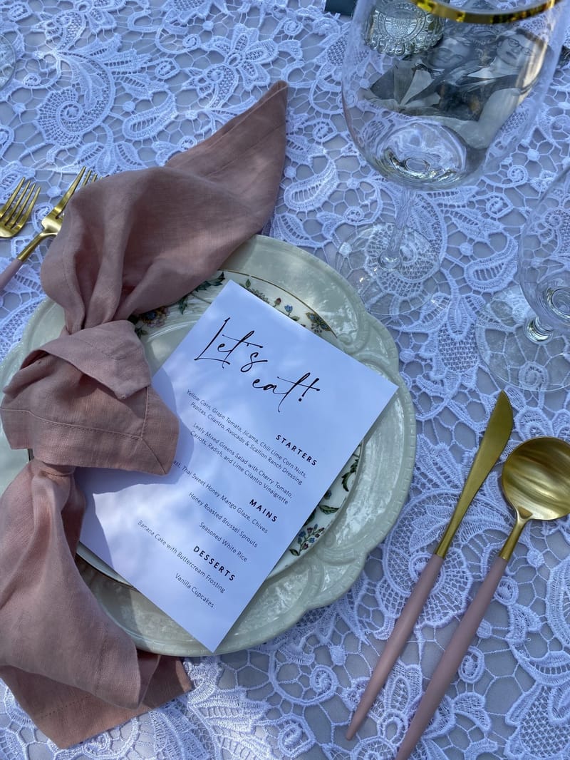 Let’s eat! menu on a floral plate with a knotted pink napkin and pink handled gold cutlery on lace